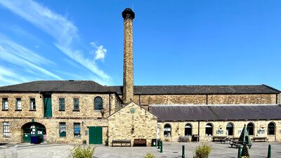 A historic brick building complex with green doors and windows stands under a clear blue sky. At its center, a tall chimney rises prominently. There are picnic tables and a few plants scattered in the courtyard area.