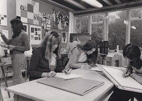 Three students sketching at a desk with two others painting at easels.